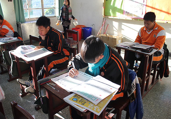 教室內，穿著橙黑色制服的學生坐在課桌前專心寫字。前景學生低頭書寫，左側學生閱讀教材，右側學生也在做功課。背景有其他學生和老師。陽光透過窗戶灑入明亮的教室。