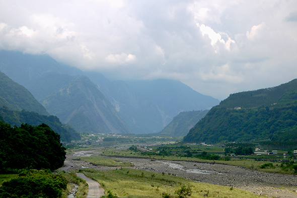 壯闊山谷風景，河流蜿蜒流經綠色平原，兩側高聳翠綠山脈環繞，山上雲霧繚繞，谷底散落民居與農田，展現原始自然的山地地形。