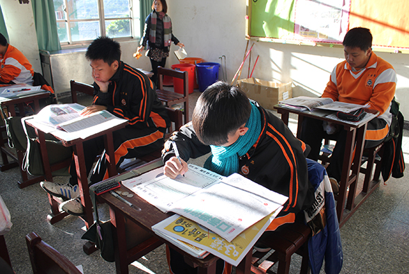 明亮的教室內，穿著橙色運動服的學生坐在課桌前專注地做功課。前景中一名學生低頭書寫，身旁放著教科書。背景還有其他學生和站立的老師。陽光透過窗戶灑入教室。