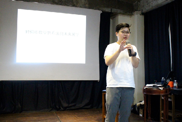 一位穿著白色短袖上衣的男性講者在室內舞台上進行演講，手持麥克風，身後投影幕上顯示日文簡報內容。舞台右側擺放有講台和設備，黑色布幕作為背景。