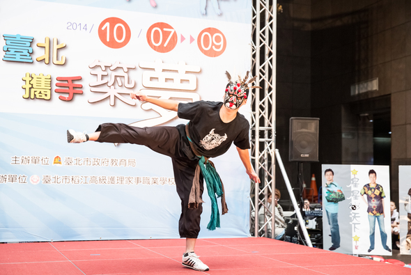 2014年北臺灣手鐵人賽事現場，一位參賽者戴著紅色面具在紅色舞台上表演高難度的單腳平衡動作，展示其體操技巧。背景標示活動日期為10月7日至9日。