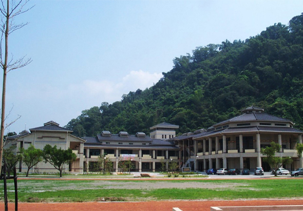 現代建築校園景觀，草坪前方有多棟白色和深色屋頂的教學建築，背景為青山翠林，晴朗天空，停放數輛車輛，環境整潔開闊。