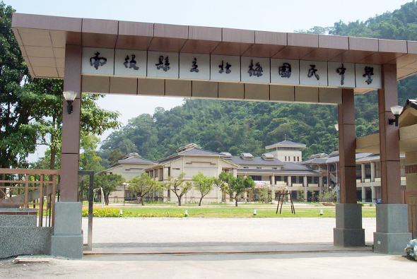 棕色木製牌坊，上方掛著十個黑色書法漢字。透過牌坊可見寬闊的校園廣場，背景為綠樹山林，右側有現代教學建築物。典型的學校入口門坊景象。