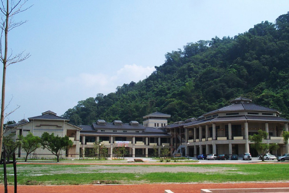 現代化校園建築群，前方為寬闊綠色草坪和紅色跑道，背景為茂密山林。建築群包括多棟淺色建築，採用傳統與現代風格混合設計，配有寬敞的走廊和柱廊。停車場停放數輛汽車，左側有裸露樹木。