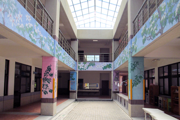現代美術館內部空間，中央走廊兩側有粉紅和淺藍色展示牆面，牆上裝飾有植物圖案藝術作品。天花板為玻璃採光天窗，兩側走廊各有黑色門窗和展示空間，地面鋪設紅磚。空間明亮寬敞，展現當代藝術展覽環境。