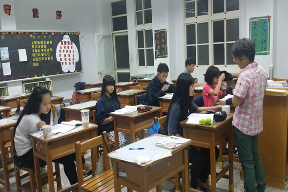 教室內，一位老師正向坐在木製課桌前的多名學生進行教學。學生們在紙上寫字或做功課，黑板上有教學內容展示，牆上貼有學習海報和裝飾品，窗戶採光充足。