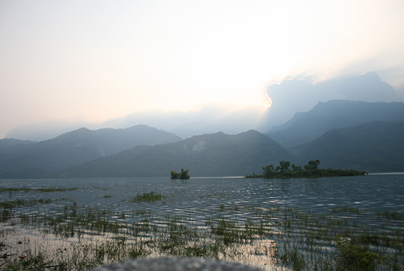 晨霧籠罩的山水風景，遠處高聳的山峰在雲霧中若隱若現，湖面平靜如鏡，近處水草茂密，中景有小島點綴，天空泛起淡淡的金黃色晨光。