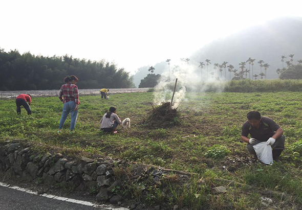 農民們在田間進行農業活動，中央燃燒著農作物殘留物，濃煙升起。幾位農民分散在田地中工作，遠景可見樹林和山脈。這是傳統農業焚燒稻草的田間勞作場景。