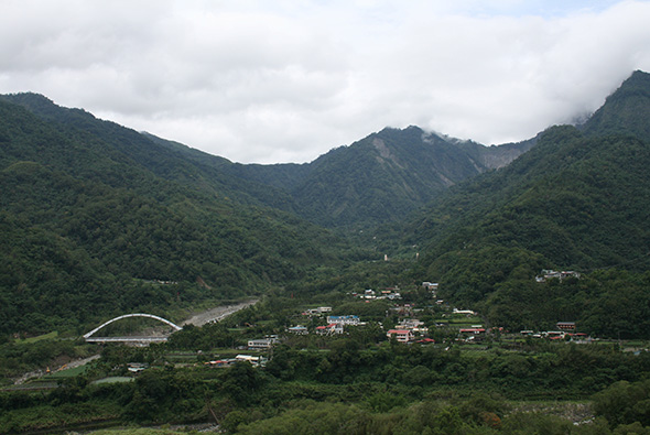 翠綠山谷中的村落景觀，密集的森林覆蓋兩側高山，山谷底部有河流蜿蜒流過，白色拱橋跨越河面，房舍聚集在河畔平地，遠處山峰籠罩於雲霧中，呈現典型的臺灣山區風貌。