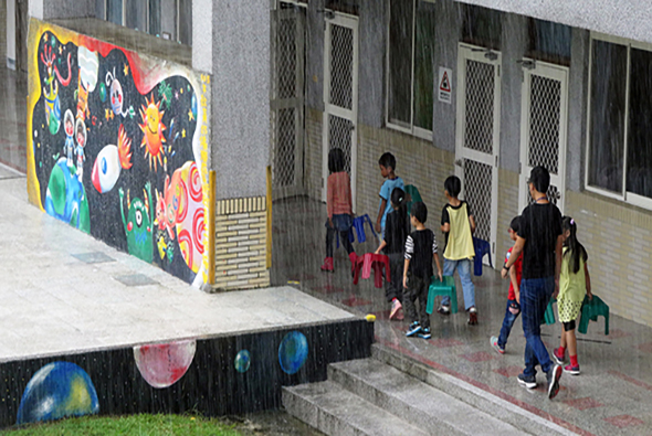 學校校園內，一群穿著校服的學童在下雨天聚集於建築物入口處。左側牆面有色彩繽紛的宇宙主題壁畫，描繪行星、星星等天體。地面有行星圖案裝飾。孩子們撐著雨傘，似乎正準備進入校舍或等候集合。