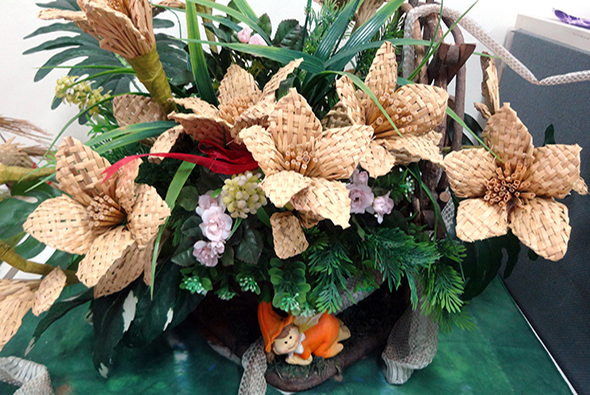 一個綠色陶土花盆，盛放著棕色網格紋理的大型乾燥花朵、紫色小花、綠色蕨類植物和紅色花材。花盆底部裝飾著小陶土動物和橙色配件，展現精心設計的乾燥花藝術作品。