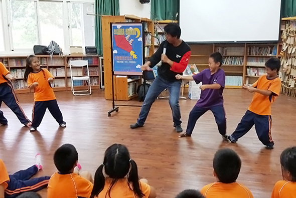 學校體育館內，一位男性教練穿著紅黑相間衣服與穿著橙色制服的學童進行互動式運動教學，多名學生圍成一圈參與活動，後排有圖書館書架及展示海報。
