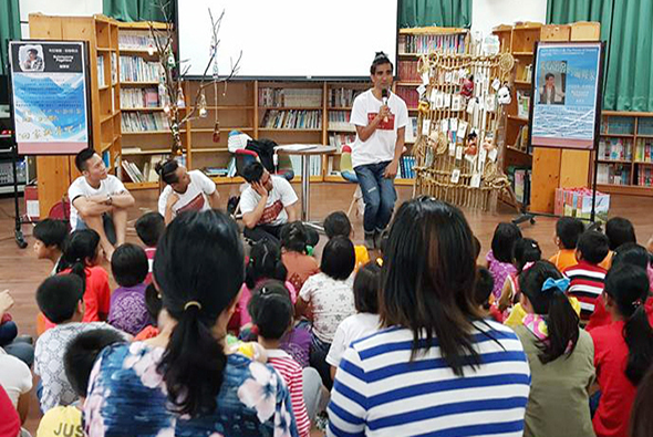 學校圖書館內，一位女性講者站在眾多小學生面前進行演講。學生們坐在地板上聆聽，身穿校服。背景是書櫃、展示板和裝飾品。這是一場校園閱讀推廣或教育活動。