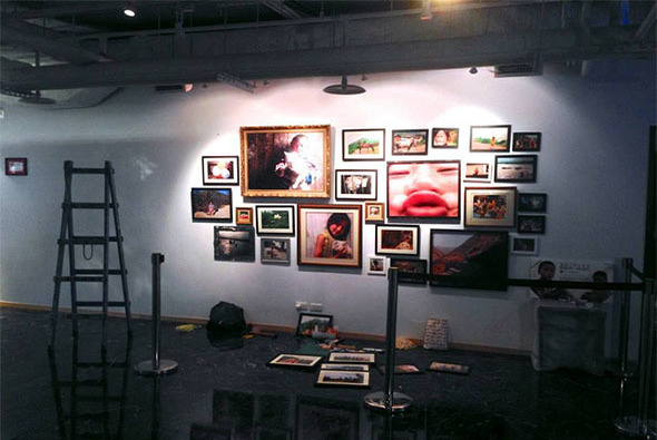 藝廊展覽現場，白色牆面上展示多幅不同風格的照片和藝術作品，包括人物肖像、風景和抽象作品。展廳地面放置梯子、展示台和其他佈置物品，燈光照亮牆面作品。
