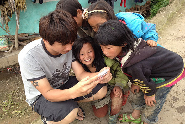 一位年輕男性蹲下與四名兒童親密互動，在戶外泥土地上一起觀看手中物品。背景有綠色建築物和其他人物。場景溫馨，展現跨代交流與關懷。
