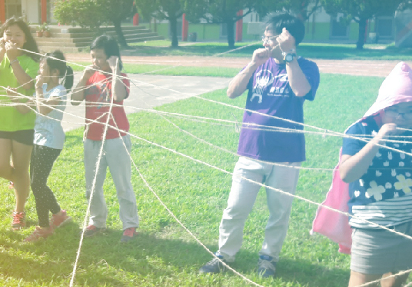 戶外公園草地上，一群兒童圍成圈，用繩子進行團隊遊戲。孩子們穿著色彩繽紛的衣服，認真參與互動活動。背景可見樹木和運動場設施，陽光灑落草地，營造歡樂的集體活動氛圍。