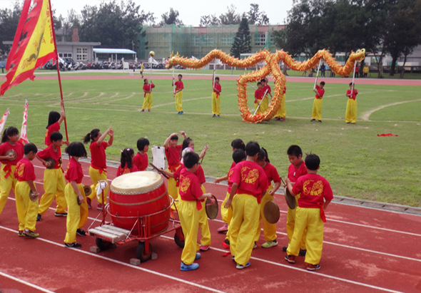 運動會開幕式現場，穿著紅黃相間服裝的表演者在田徑場進行舞獅表演。舞台上擺放著大型金色舞獅道具，場邊豎立紅黃相間的旗幟，觀眾席坐滿人群，背景為校舍建築。