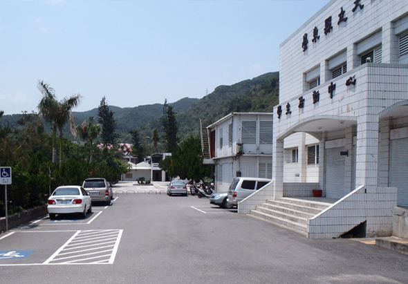醫院停車場場景，停放數輛白色汽車，右側為白色磁磚現代醫院建築，前方有樓梯和入口，背景為青翠山丘和綠色樹木，天氣晴朗。