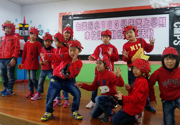 學校禮堂內，約十五位穿著紅色服裝和紅帽的小學生在舞台上進行表演活動。背景黑板上有中文標題，舞台設有彩色布景。孩子們擺著各種動作姿勢，看起來在進行團體舞蹈或才藝展演。