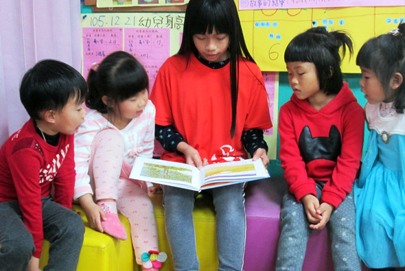 幼兒園教室內，一位穿著紅色上衣的老師坐在黃色座位上，為五名穿著紅色衣物的小朋友講述繪本故事。孩子們圍坐在老師身邊專注聆聽，教室背景有彩色教學海報和學習用品。