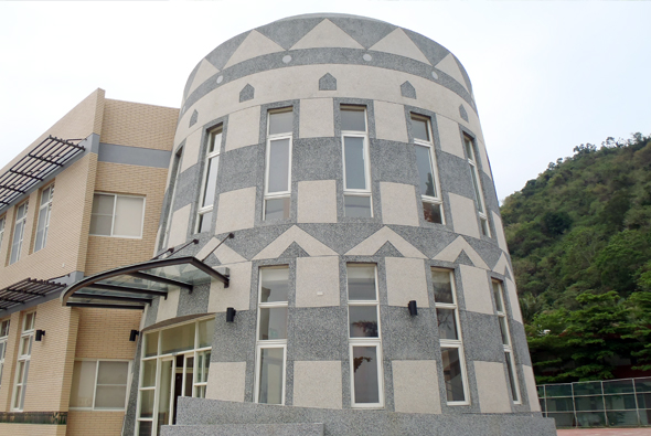 圓筒形現代建築物，外牆採米色和灰色方塊交錯拼貼設計，頂部為幾何三角形屋頂。建築配備多扇大型窗戶，左側為米色方形建築，背景為青山綠樹。