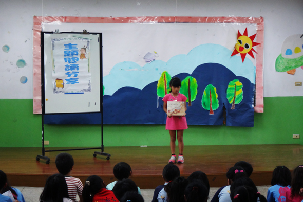 小學教室內，一名穿著紅色衣服的學生站在舞台上舉著書本進行讀書分享。舞台背景有藍色山丘、綠色樹木和黃色太陽的彩繪裝飾。台下坐著多名學生觀看。左側豎立著展示書籍介紹的黑色立架。