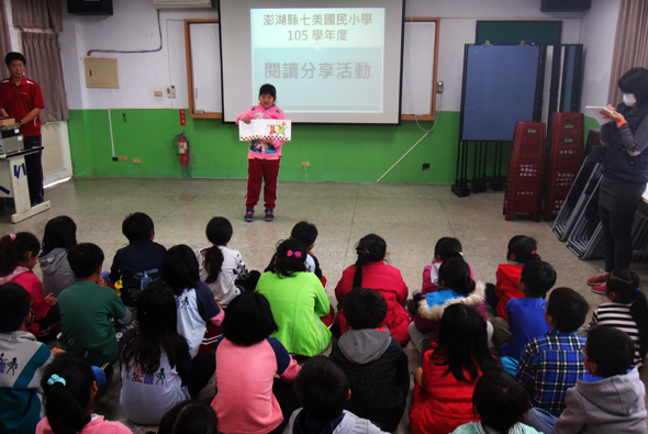 港澳慈心國小105學年度閱讀分享活動，學生聚集在綠色運動地板的室內場地，一名穿著紅白相間服裝的學生站在講台前進行分享，其他同學坐在地上認真聆聽，牆上投影幕顯示活動標題。