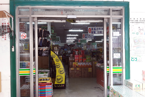 一間便利商店的店面，綠色框架的玻璃門窗，門口擺放黃色促銷海報和彩色堆疊商品。店內可見貨架、商品陳列和日光燈照明。門邊貼有公告和標誌。