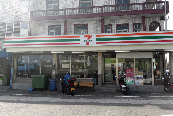 一間7-Eleven便利店的外觀，位於街道底層，紅白綠色招牌明顯，玻璃窗展示店內商品，門前停放數輛機車，樓上為民宅建築，白色瓷磚牆面搭配木製陽台。