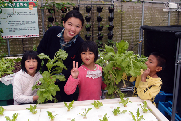 一位女性教師與四名小學生在室內垂直農場合影。背景有懸掛的植物種植裝置和教育標語牌。學生們手持剛採收的生菜等蔬菜，展示農業教育成果，洋溢歡樂的學習氛圍。