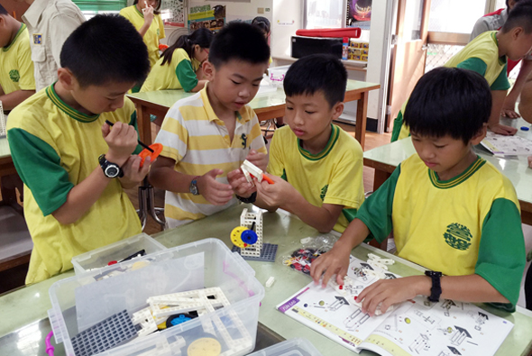 一群穿著黃綠色制服的小學生在教室內進行科學實驗活動。學生們圍坐在桌邊，正在組裝或操作機器人模型和電子元件。桌上擺放著各種實驗工具、零件和教學材料。背景可見其他學生也在進行類似的STEM學習活動。