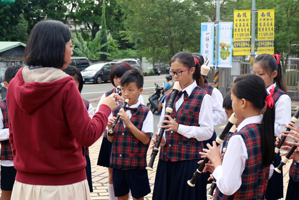戶外音樂表演活動，一位女性主持人手持麥克風與穿著紅格紋背心制服的學童進行互動。多名學生手持木笛樂器，樹木和停車場可見於背景，黃色宣傳旗幟豎立在現場。