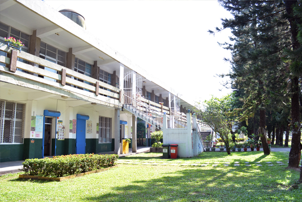一棟兩層樓的學校建築，白色牆面配藍色門窗，前方有綠色草坪和灌木叢。建築右側立有高大樹木，左側可見樓梯和欄杆。校園環境寧靜整潔。