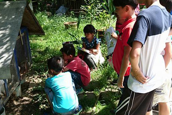 一群兒童和青少年在戶外草地上圍成一圈，參與某項集體活動或遊戲。穿著紅色、藍色和灰白色衣服的參與者專注地看著圓圈中央，背景可見綠色植被和遮陰處。