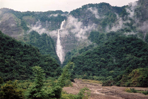 高聳的瀑布從雲霧繚繞的陡峭山崖傾瀉而下，周圍被茂密的綠色森林包圍。山谷底部有河流和裸露的岩石地表，展現出壯觀的山區自然景觀。