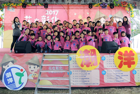 2017年花在彰化活動舞臺上，大約五十位穿著粉紅色上衣的兒童和工作人員合影。舞臺背景為粉紅黃色設計，展示活動海報、時程表和彰化縣徽標誌。