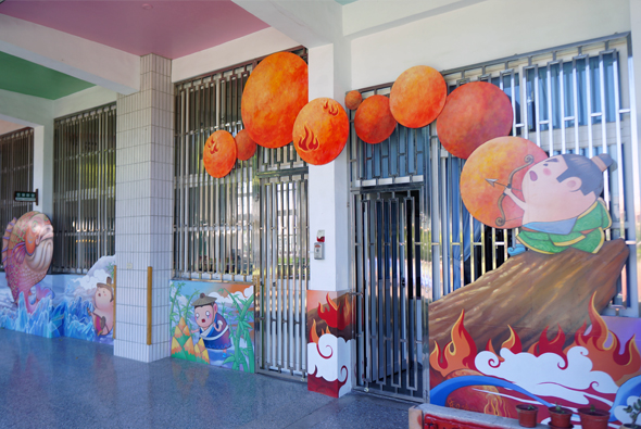 幼兒園教室內，天花板懸掛橙紅色紙質太陽裝飾，牆上繪有卡通人物和火焰圖案。鐵欄門前擺放彩色壁畫，展現兒童藝術創作主題。室內採光明亮，佈置溫馨童趣。