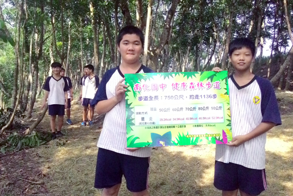 兩名穿著學生制服的學生在樹林中舉著綠色宣傳海報，海報上有中文文字和活動資訊。背景可見其他學生在林間活動。這是一場校園環保或健康推廣活動的宣傳照片。