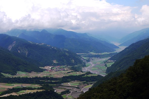 壯麗的高山谷地風景，俯瞰綠色農田和蜿蜒河流穿過寬闊山谷，遠處層疊青山在白雲籠罩下延伸至天際，呈現典型的阿爾卑斯山區地貌。