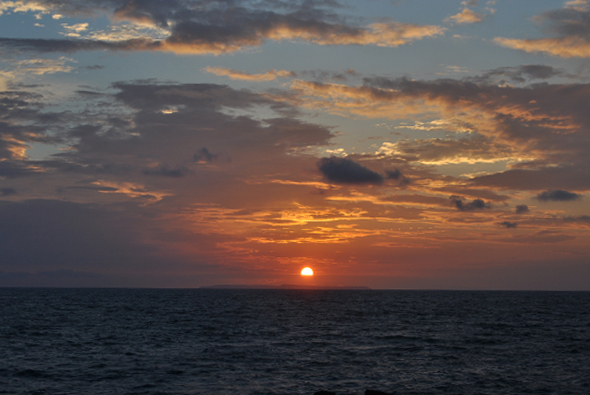 夕陽西下，金黃色的太陽逐漸沉入海平線，天空被染成橙紅色和深藍色的漸層。厚重的烏雲被夕陽照亮，呈現金色邊緣。平靜的海面映照著落日光芒，營造出寧靜祥和的黃昏景象。