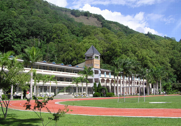 一所現代化學校建築坐落在青翠山麓下，白色多層校舍配備紅色跑道運動場，綠草坪中央，棕櫚樹點綴校園，背景為密集的綠色森林覆蓋的山丘，藍天白雲映照。