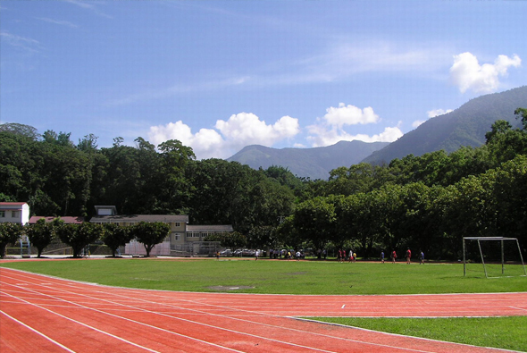 運動場景：紅色跑道環繞綠色草坪，遠處可見足球門。背景是茂密的樹林和高聳的山脈，天空晴朗有白雲。左側建築物和看台可容納觀眾，多人在草地上活動，呈現戶外運動設施的風貌。