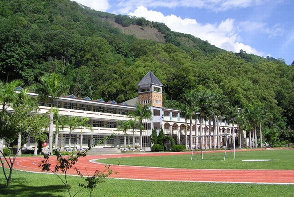 一座現代化白色建築坐落於綠山山腳，配有尖塔和棕櫚樹。前方為紅色跑道和寬闊草坪，背景為密集的綠色山林，晴朗天空中飄著白雲。