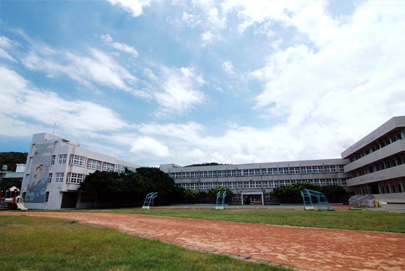 校園操場全景，紅土跑道圍繞綠色草坪，四周環繞現代校舍建築，晴空萬里，白雲朵朵，操場上設有運動設施，是學生進行體育活動的場所。