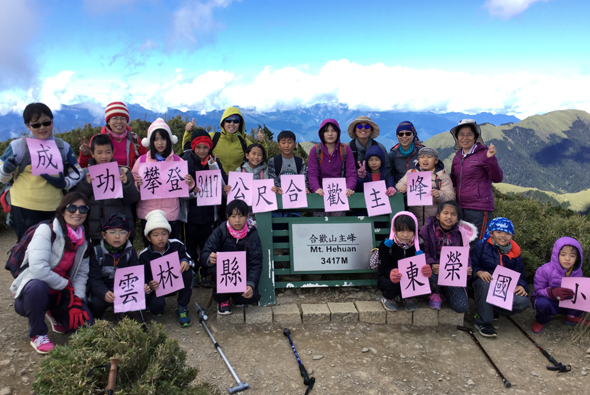 登山隊伍在海拔3417公尺的合歡山主峰山頂，集體舉著粉紅色紙張排成漢字，背景是壯闊的山脈和藍天白雲，眾人穿著登山服裝歡樂地合影留念。