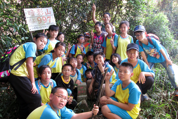 一群穿著黃藍相間隊服的登山隊員在山林中合照，舉著標示「唧影山」的牌子，背景是茂密的樹木。隊伍包含男性和女性登山者，展現團隊精神，記錄登山活動的成果。