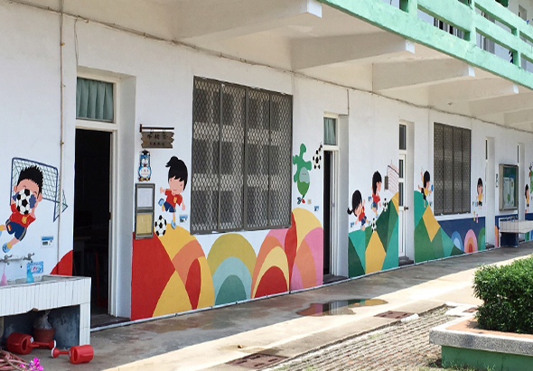 幼兒園走廊外牆繪有彩色壁畫，包括紅、黃、綠、藍等鮮豔色塊和卡通人物圖案。走廊沿線設有多扇門窗和通風口。左側放置紅色玩具和植物盆栽，營造溫暖的兒童學習環境。