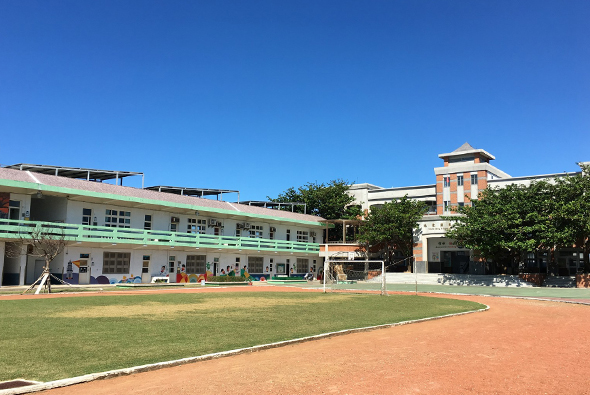 學校運動場景，前景為紅土棒球場，中間是綠色草坪，後方為綠色和橙色建築物組成的校舍，晴朗藍天，場地整潔有序，適合體育教學和運動比賽。