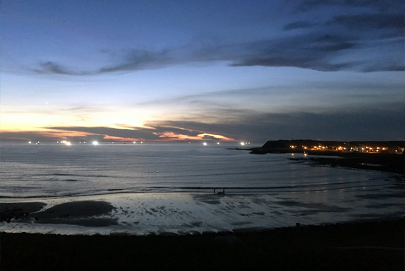 黃昏時分的海灘景色，天空呈現藍紫色漸層，地平線上有橙黃色的落日餘暉。右側遠處山丘上點綴著城鎮燈火。前景為黑色沙灘和輕柔的海浪。