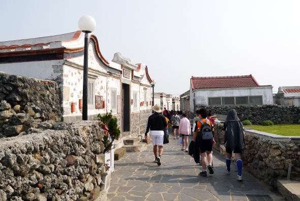 遊客在中式古建築前的石板路上漫步，建築具有傳統的翹角屋頂和白牆設計。周圍環繞著古老的石牆，遠處可見紅瓦屋頂的建築物和綠色田野。